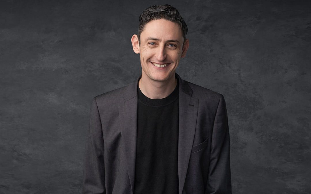 A professional man smiling wearing a grey suit and black Tshirt