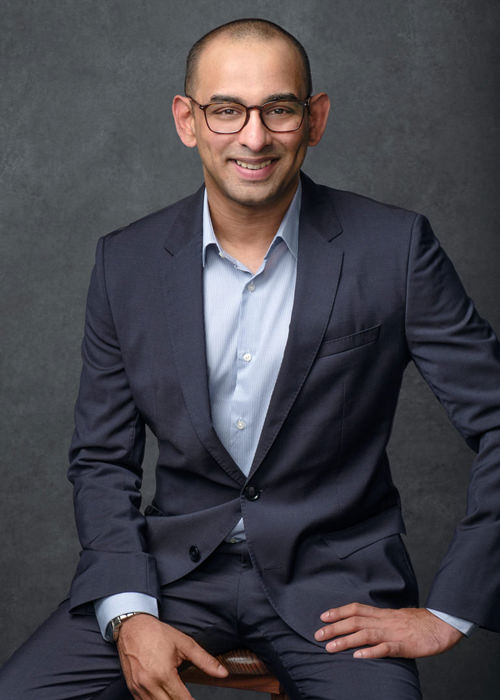 A professional portrait of a young Indian businessman sitting down and smiling wearing a suit